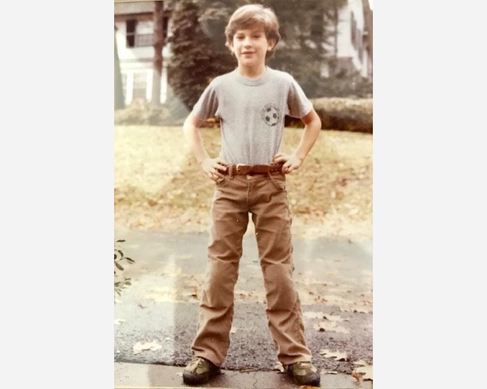 Dan Harris as a child standing in a driveway with his hands on his hips. He is wearing a grey T-shirt tucked into brown corduroy trousers.